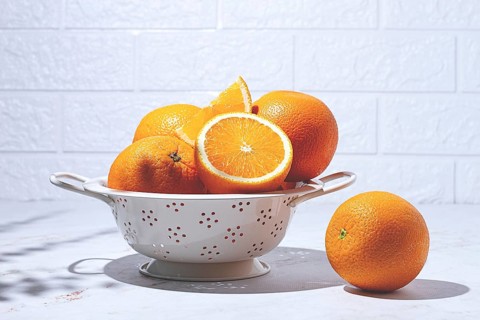 Oranges are resting in a white colander on a light surface. One orange sits next to the colander