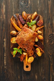 Roasted pork meat on wooden board surrounded by roasted potatoes, carrots and beets. Board is on