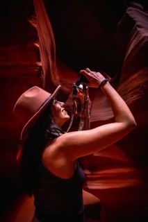 Woman wearing a hat and black tank top, smiling and looking up, holding a camera to photograph the