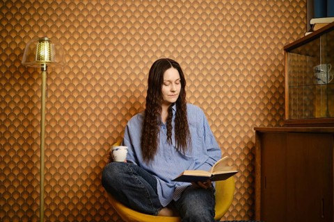 Woman sits in yellow chair, reading book while holding cup. Room has brown patterned wallpaper and