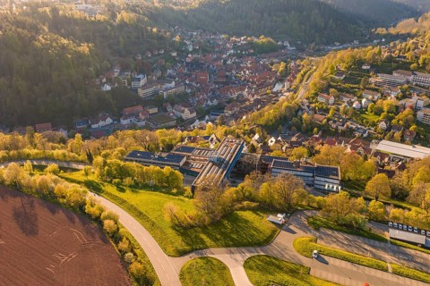 Aerial view of a village in a hilly, green landscape, roads and fields visible in sunshine, Calw