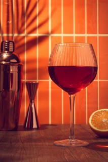 A drink is placed on a wooden table. A metal shaker and measuring cup stand nearby. Orange tiles