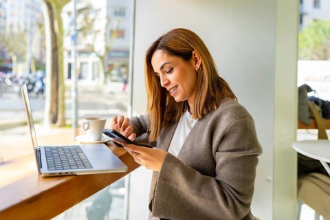 Young woman working remotely in a modern cafe, smiling as she scrolls and taps her smartphone