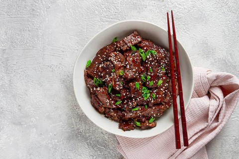 Crispy beef, with green onions and sesame seeds, in a bowl with chopsticks, on a textured surface,