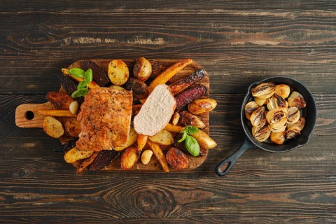 Roasted pork loin on wooden board surrounded by various vegetables. A small pan holds cooked onions