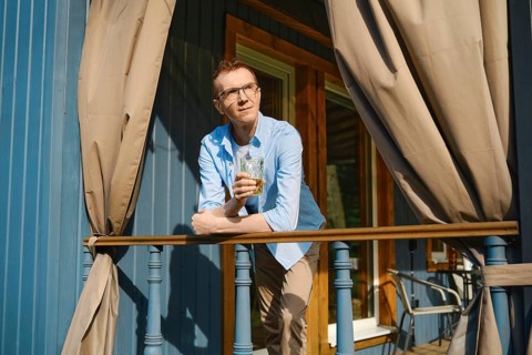 Middle-aged man in blue shirt and beige trousers stands on terrace of wooden cabin looking