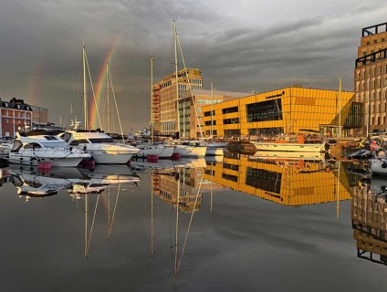 Modern buildings and boats are reflected in harbor water under a dramatic sky with rainbow,