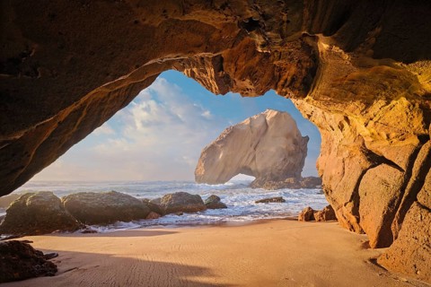 Sunset casts warm colors over Penedo do Guincho rock arch with waves crashing on sandy beach,