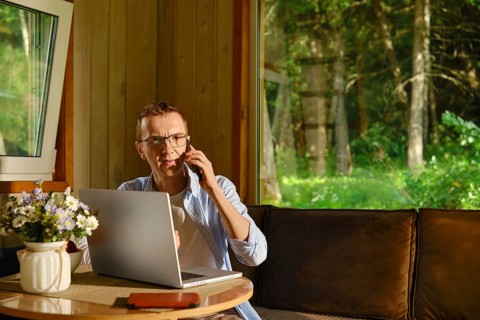 Middle-aged man sits at table in wooden cabin, talking on phone while using laptop. He solves