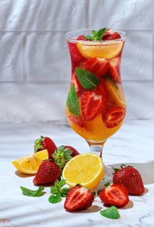 A tall glass filled with a colorful drink made from strawberries, lemon slices, and mint leaves.