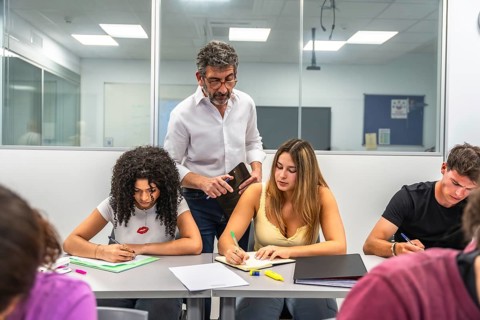 Teacher standing over students, helping with a test while they focus intently on writing answers,