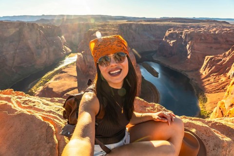 Woman wearing sunglasses and a bandana taking a selfie, sitting on a rock formation overlooking the