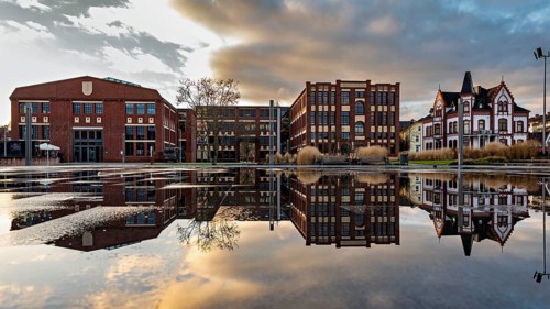 Modern and historic buildings are reflected in calm water, under a dramatic sky, The Word-Rich