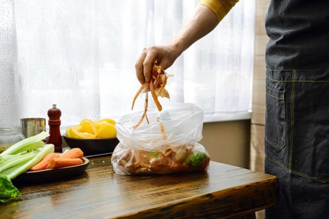 A man is standing in a home kitchen, adding vegetable scraps to a clear bag to add the waste to the