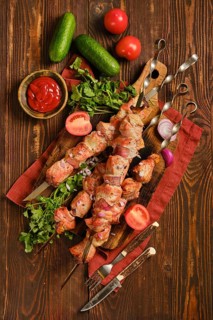 Top view of juicy shish kebab on a wooden board. Fresh tomatoes, cucumbers, and herbs surround the