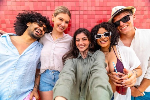 Diverse group of smiling young friends leaning against a red tiled wall, taking a selfie together,