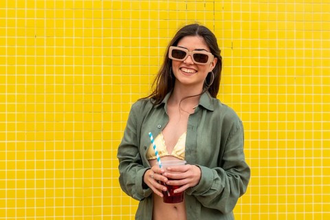 Young woman smiling, wearing sunglasses and a casual shirt over a swimsuit, holding a refreshing