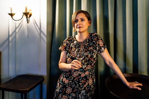 A blonde in a dress with a wine glass stands near a wine barrel. A woman in a dark floral dress