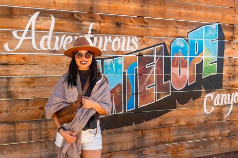 Young woman smiling and wearing a hat, sunglasses, scarf, and white shorts, standing in front of a