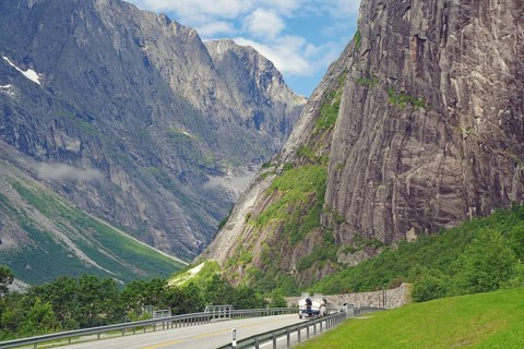 Motorcyclists on a lonely mountain road, flanked by steep cliffs and green mountains under a blue