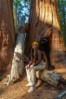 Young woman smiling, wearing a beanie and backpack, sitting on a fallen log while enjoying the