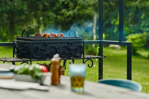Skewers are cooking on barbecue grill in garden. Smoke rises from meat. Nearby table has fresh