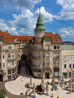 Baroque building view with green tower and red roofs surrounded by shops and cafe under cloudy sky,