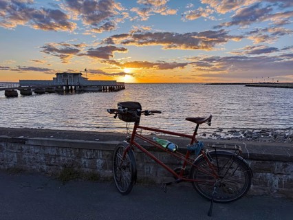 A bicycle is parked in front of a stone wall with a view of the sea at sunset. A pier stretches