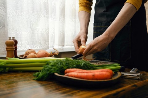 A person in apron peels an onion while standing at a kitchen counter. Fresh carrots and celery are