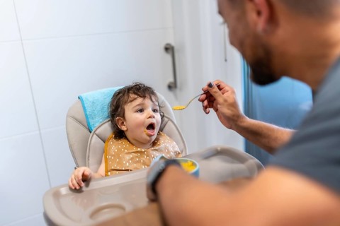 Father feeding vegetable puree to a happy baby in a high chair, celebrating family bonding and