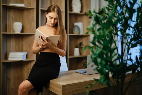 A woman sits on a desk in a modern office, reading notes. The room has wooden shelves with