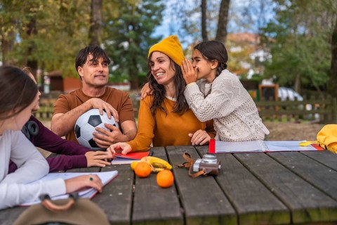 Family gathered at a wooden picnic table in a park as a daughter whispers to her mother, capturing