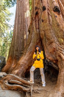 Young woman smiling, wearing a yellow jacket and carrying a camera, standing among massive ancient
