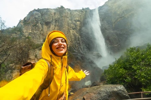 Smiling young woman exploring nature, wearing a bright yellow raincoat and beanie, happy standing