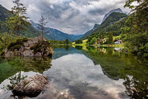 Clear mountain lake with rocks and tree in the foreground, framed by wooded alpine slopes, The