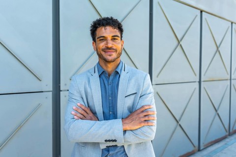 Young handsome man wearing a gray suit jacket and blue shirt, crossing arms and smiling at the