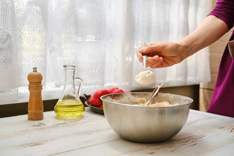 A person is measuring flour with a spoon over a mixing bowl. Fresh vegetables and cooking oil are