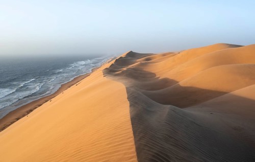 Sand dunes with wave patterns and sea in evening light, wind blowing sand across the dunes,