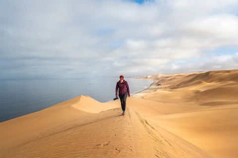 Young woman with camera standing on a sand dune in front of impressive desert landscape and sea,