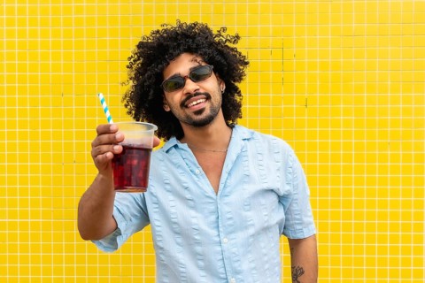 Young man with curly hair and sunglasses smiling while holding a cold drink with a straw, standing