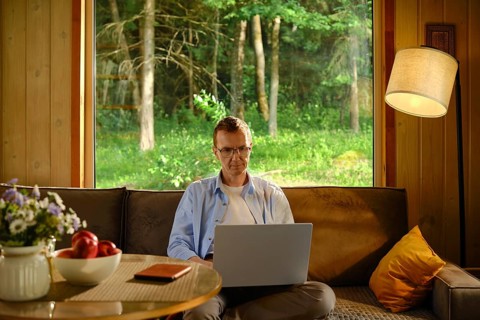 Middle-aged man sits on couch in wooden cabin. He focuses on laptop while making online shopping
