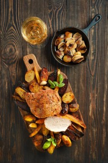 Roasted pork loin on a wooden board along with assorted vegetables. A glass of white wine sits