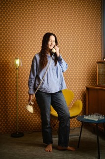 Woman stands barefoot in retro room, holding rotary phone and talking. Chess set on table and