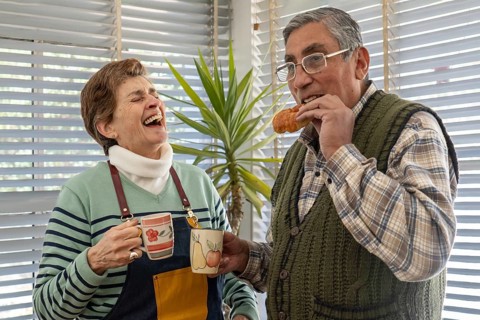 A senior woman bursts out laughing while holding two colorful ceramic mugs as her husband eats a