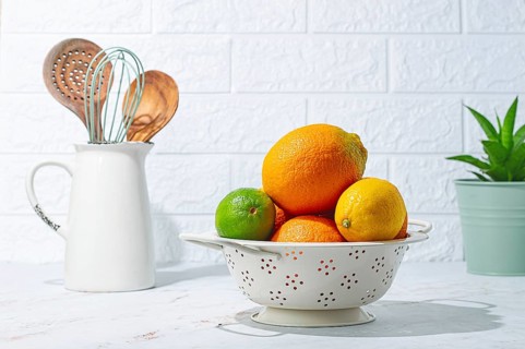 A bowl holds different fruits including oranges, lemons, and a lime on a table. Kitchen tools are