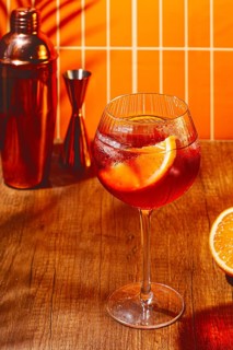 A cocktail glass filled with a drink and an orange slice sits on a wooden table. Copper bar tools