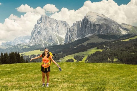 Back view middle aged gray hair woman hiker arms outstretched with yellow backpack overlooking lush