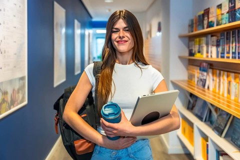 Young female student smiling while walking through a university hallway or library, carrying a