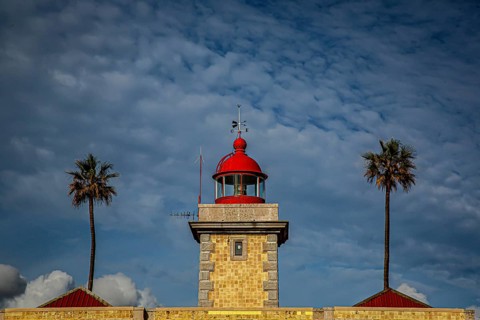 A lighthouse with red roofs and two palm trees against a cloudy, blue sky, Farol da Ponta da