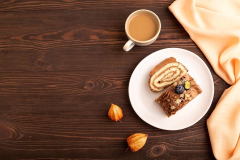 Sweet loaf with cream and caramel, cup of coffee, on brown wooden background with orange textile,